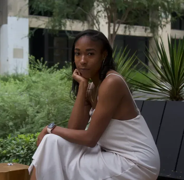 Woman in a white dress sitting on a bench in a garden, resting her chin on her hand.