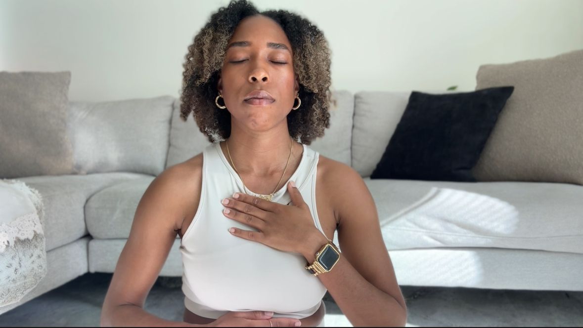Person meditating indoors on a yoga mat, eyes closed, hand on chest, in front of a gray couch.