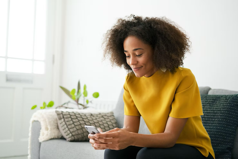 Woman in a yellow top smiling at her phone on a couch in a bright living room