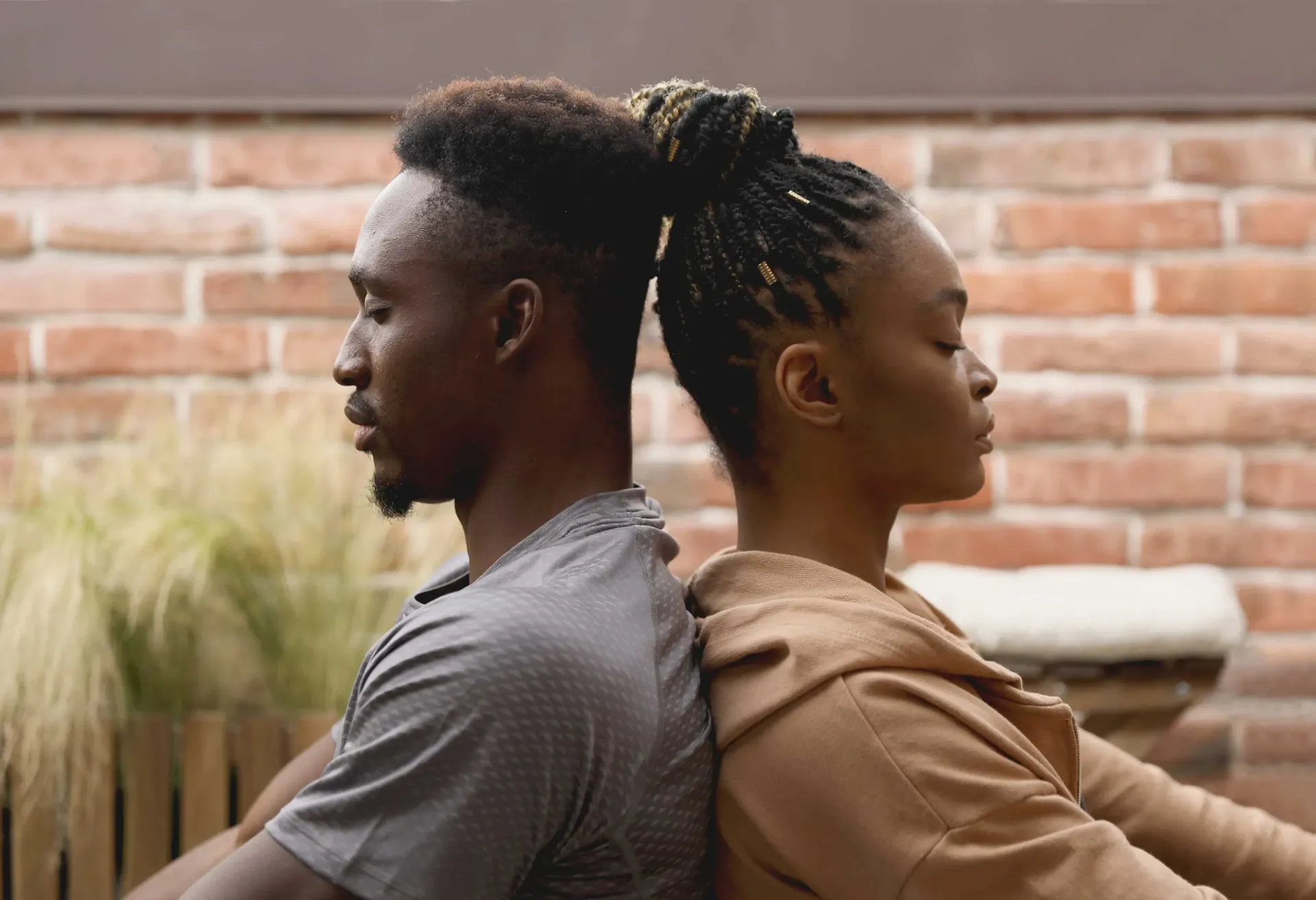 Two people sit back-to-back outdoors against a brick wall, facing opposite directions.