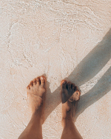 Bare feet standing on wet sand, with waves washing over them and a long shadow beside them