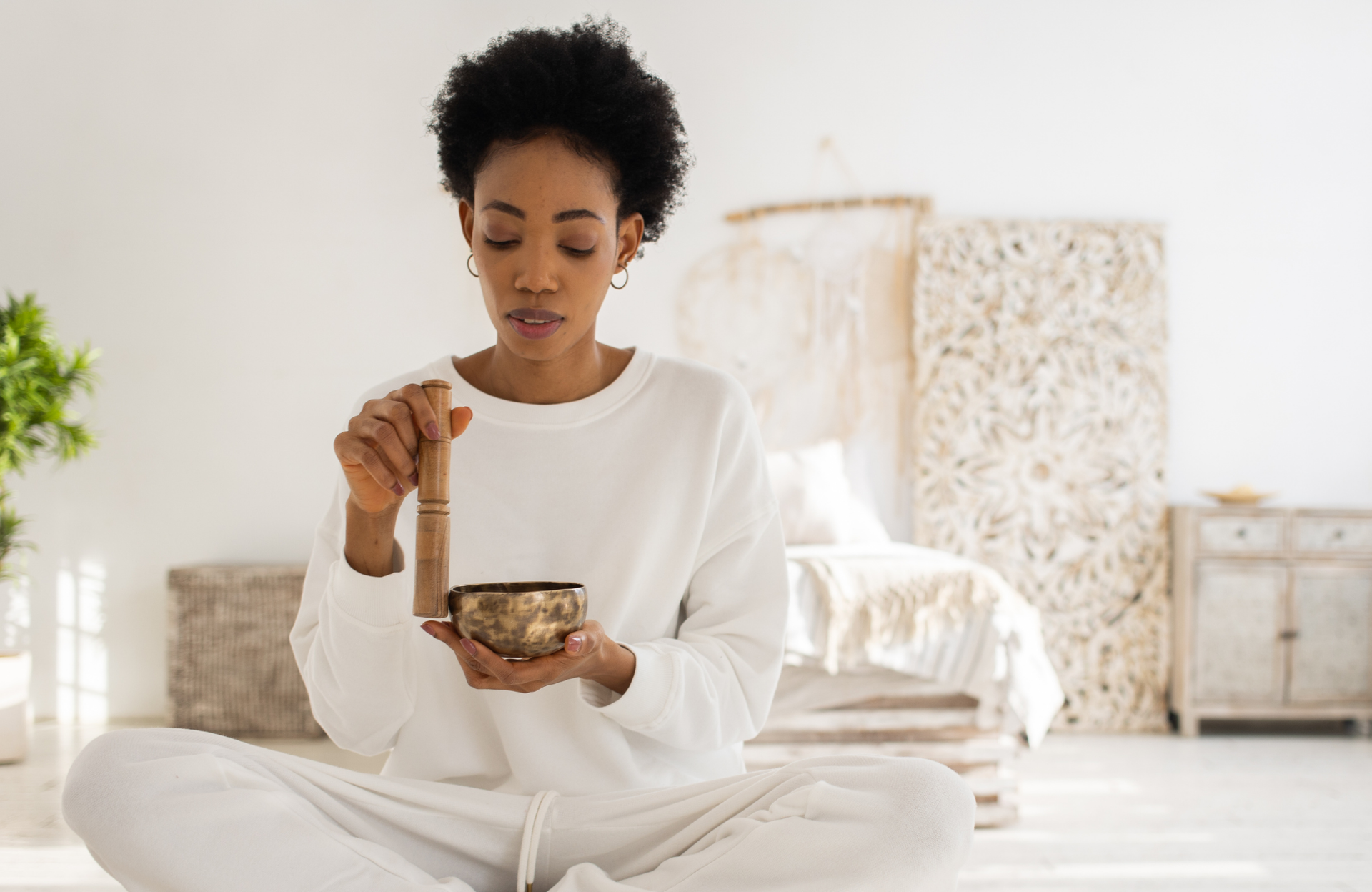 Person seated indoors, using a wooden mallet and singing bowl in a bright, minimalist room.