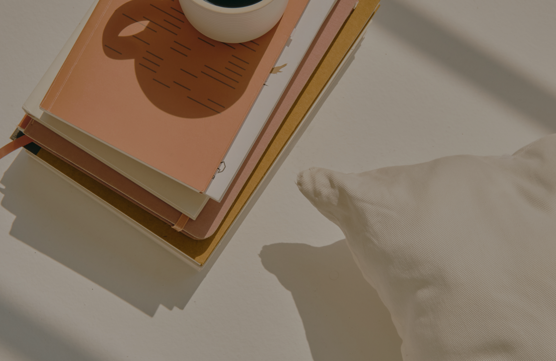 Stacked notebooks with a coffee cup on a white desk beside a white pillow