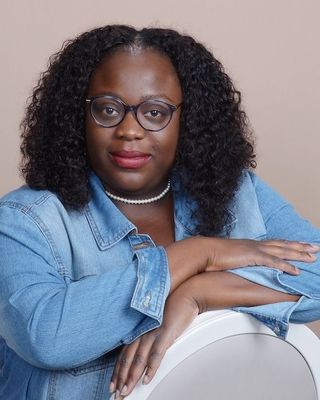Smiling person with glasses in a blue patterned sweater, standing by a yellow brick wall.