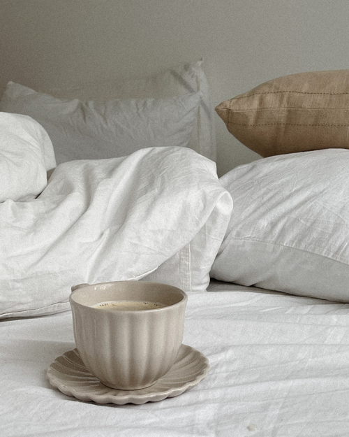 Beige teacup on a saucer on white bedding with pillows in the background