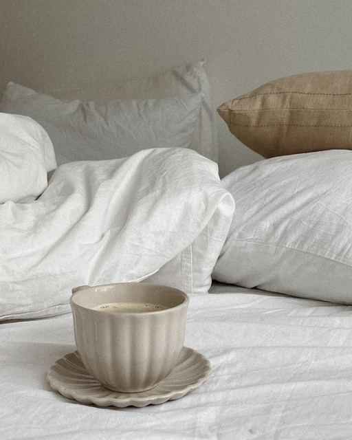 Beige teacup on a saucer on white bedding with pillows in the background