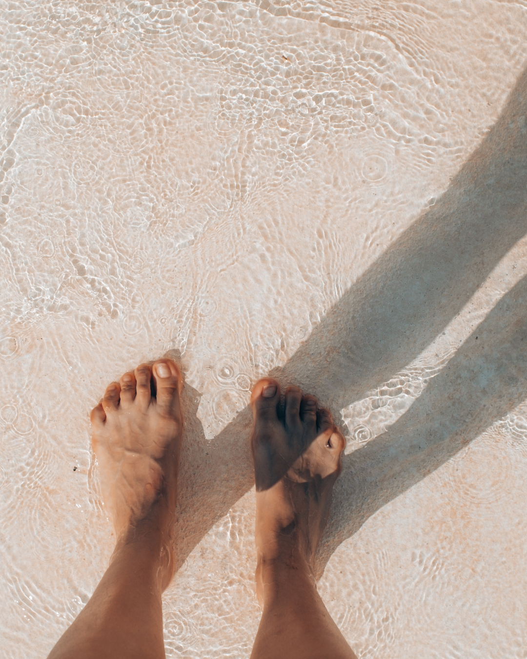 Bare feet in shallow surf on pale sand, with long shadows stretching across the beach.