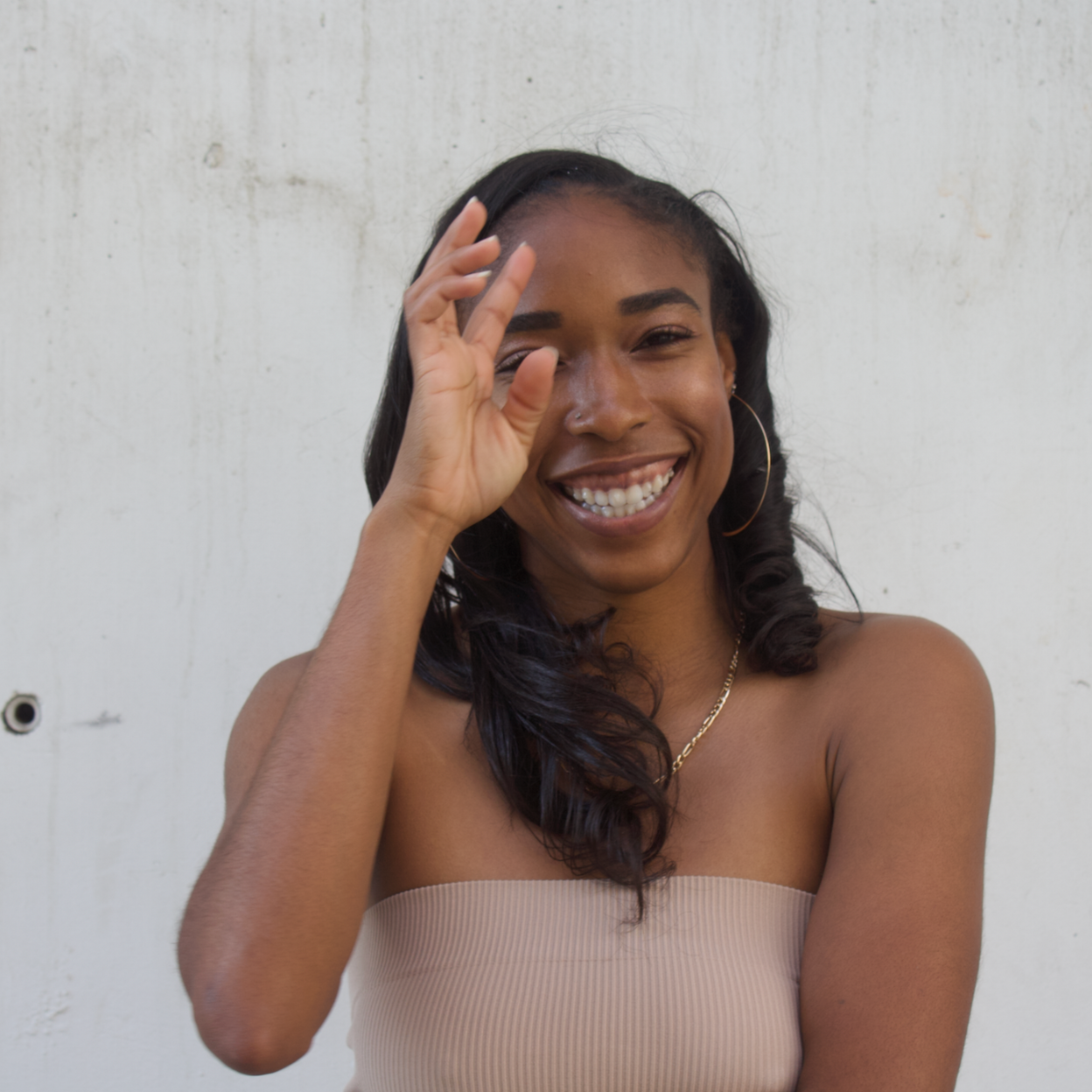 Smiling person in a strapless top, touching their face against a plain white wall.