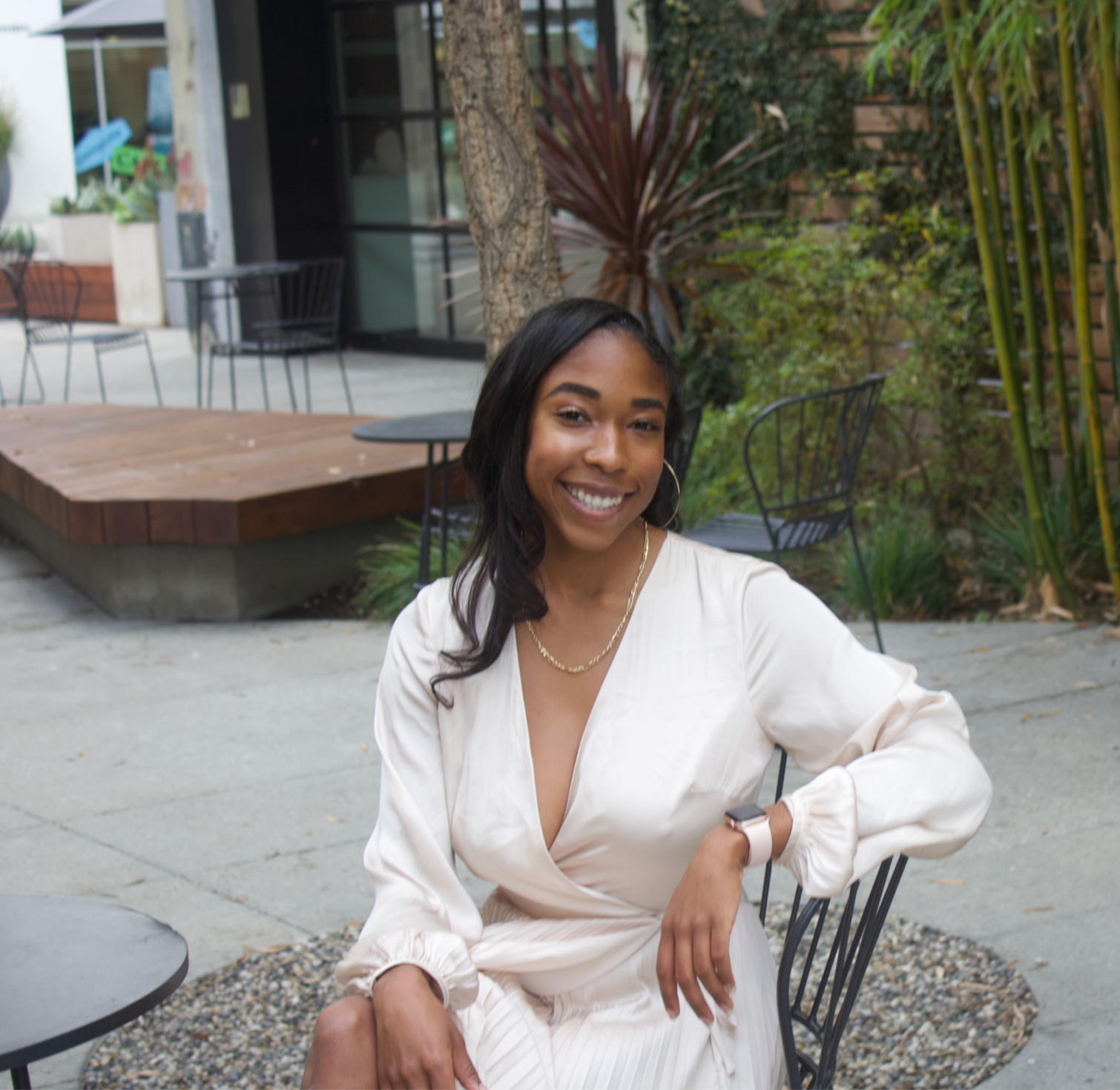 Smiling woman in a white dress seated outdoors on a patio with plants and modern seating