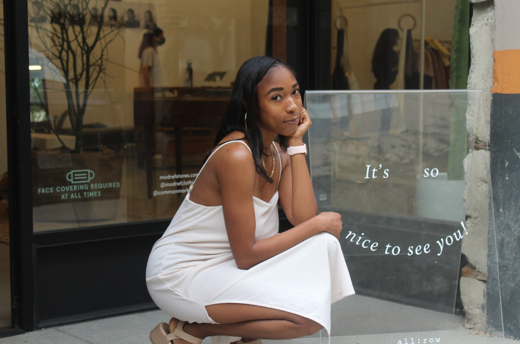 Woman in a white dress crouches on a sidewalk outside a storefront, resting her chin on her hand.