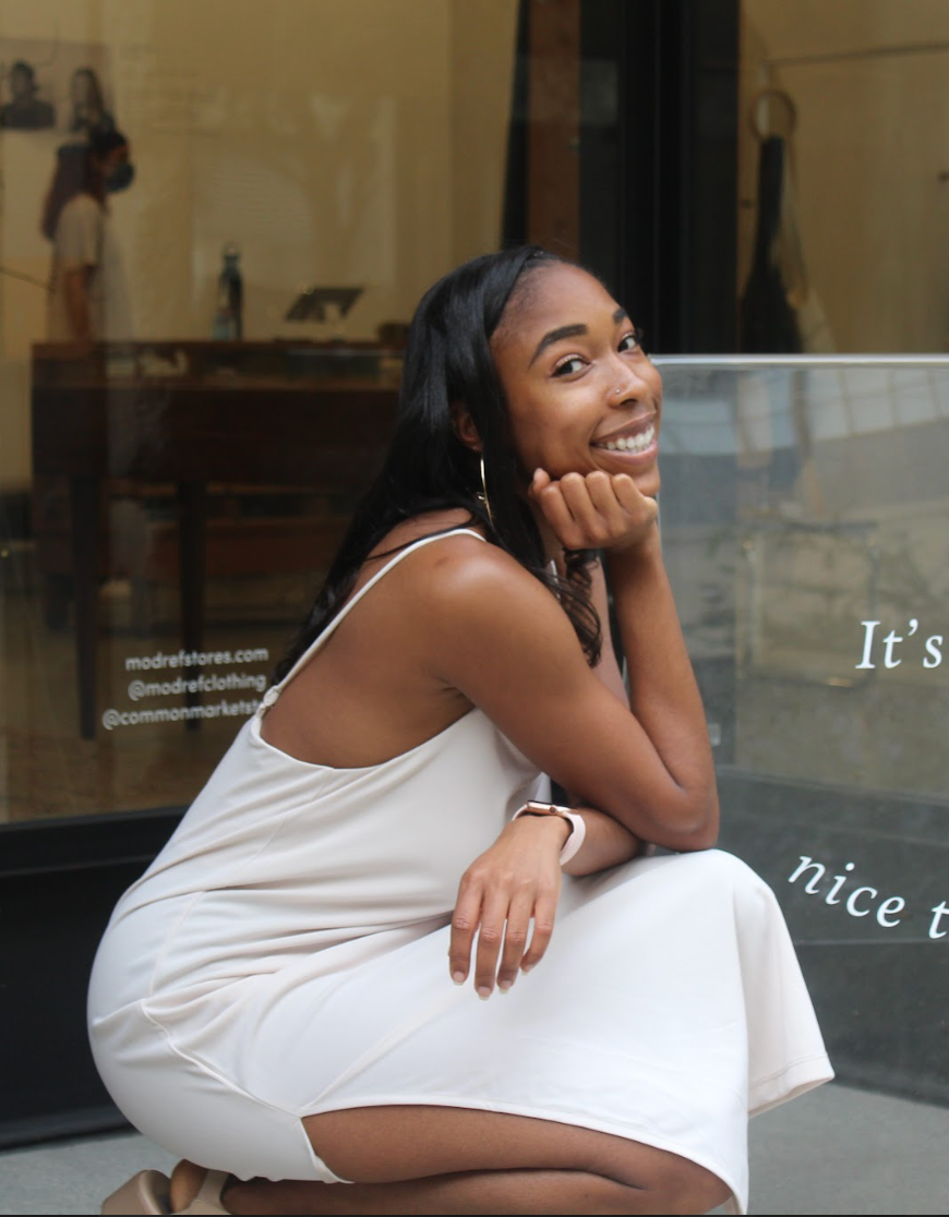 Woman in a white dress crouches and smiles outside a storefront.