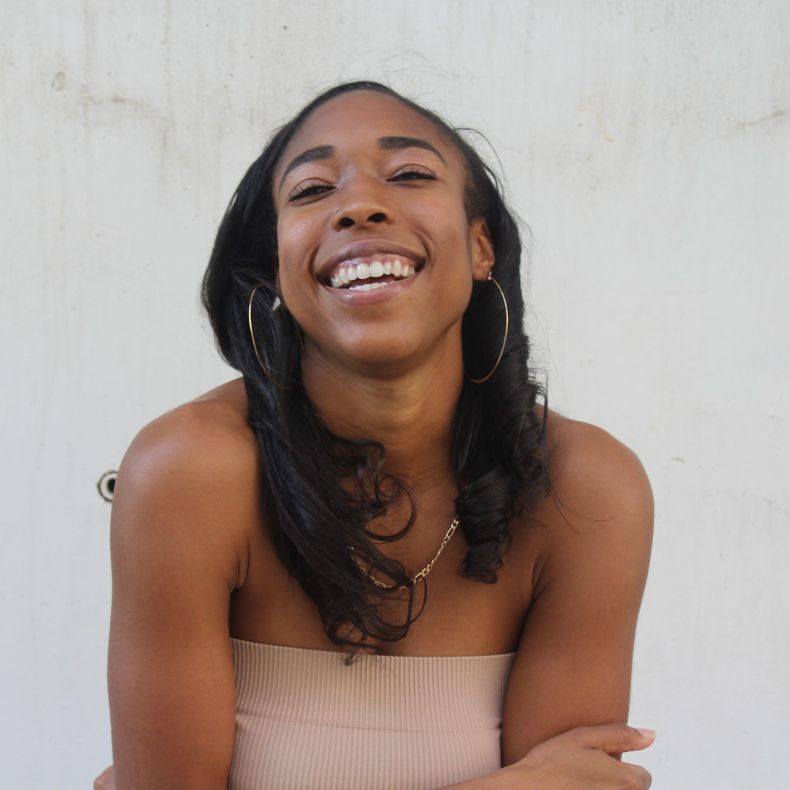 Smiling person with long dark hair and hoop earrings, wearing a strapless top against a light wall