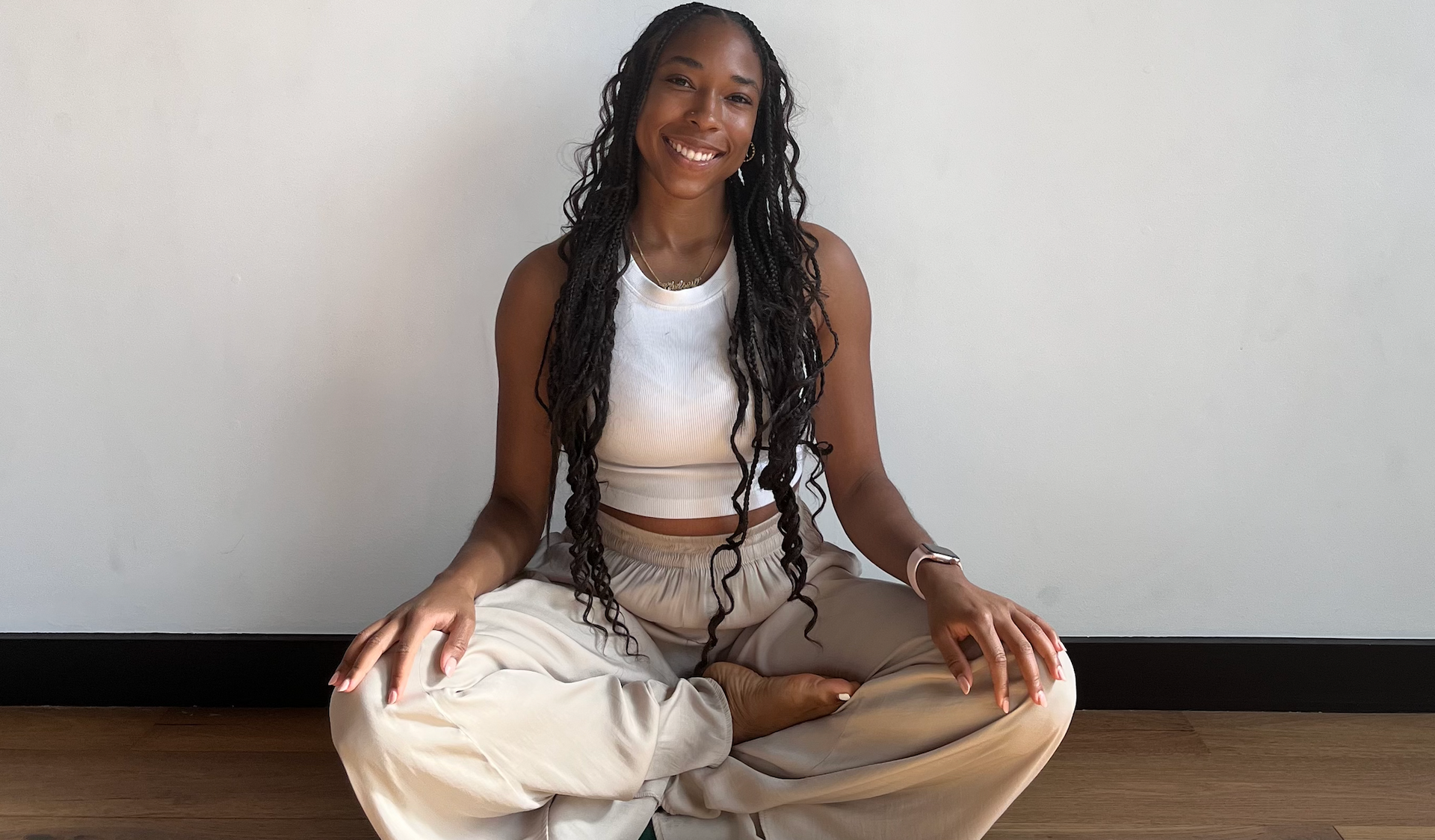 Woman sitting cross-legged on a wooden floor, smiling in a white top and beige pants.