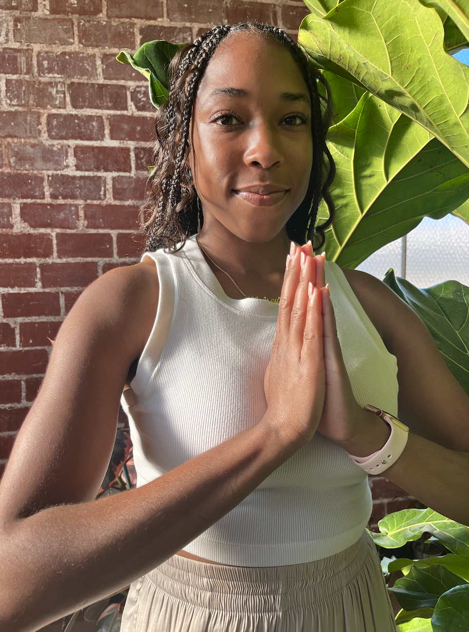 Person in white top holding prayer pose indoors beside a brick wall and large green plant