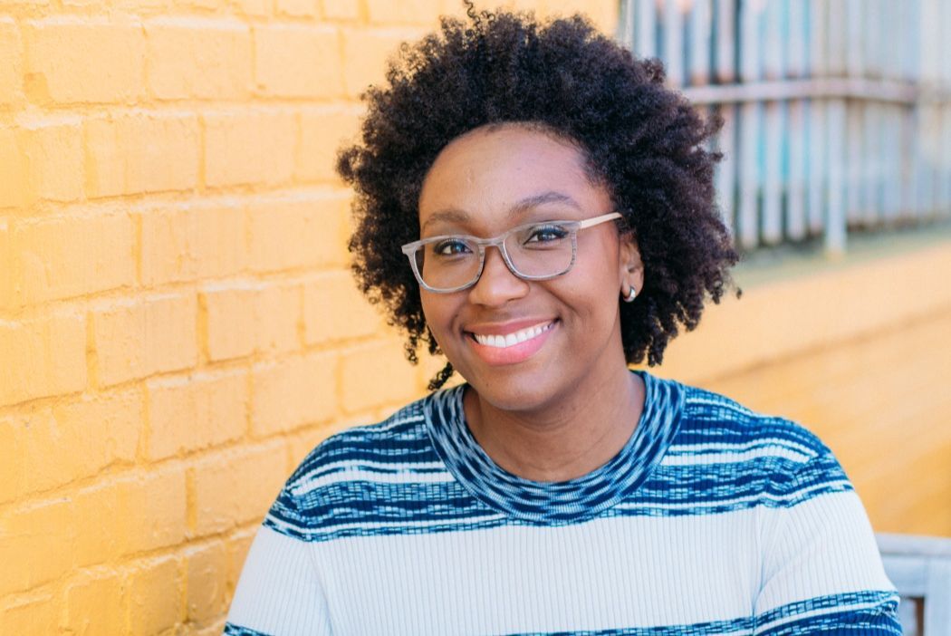 Smiling person with glasses in a striped shirt standing by a yellow brick wall