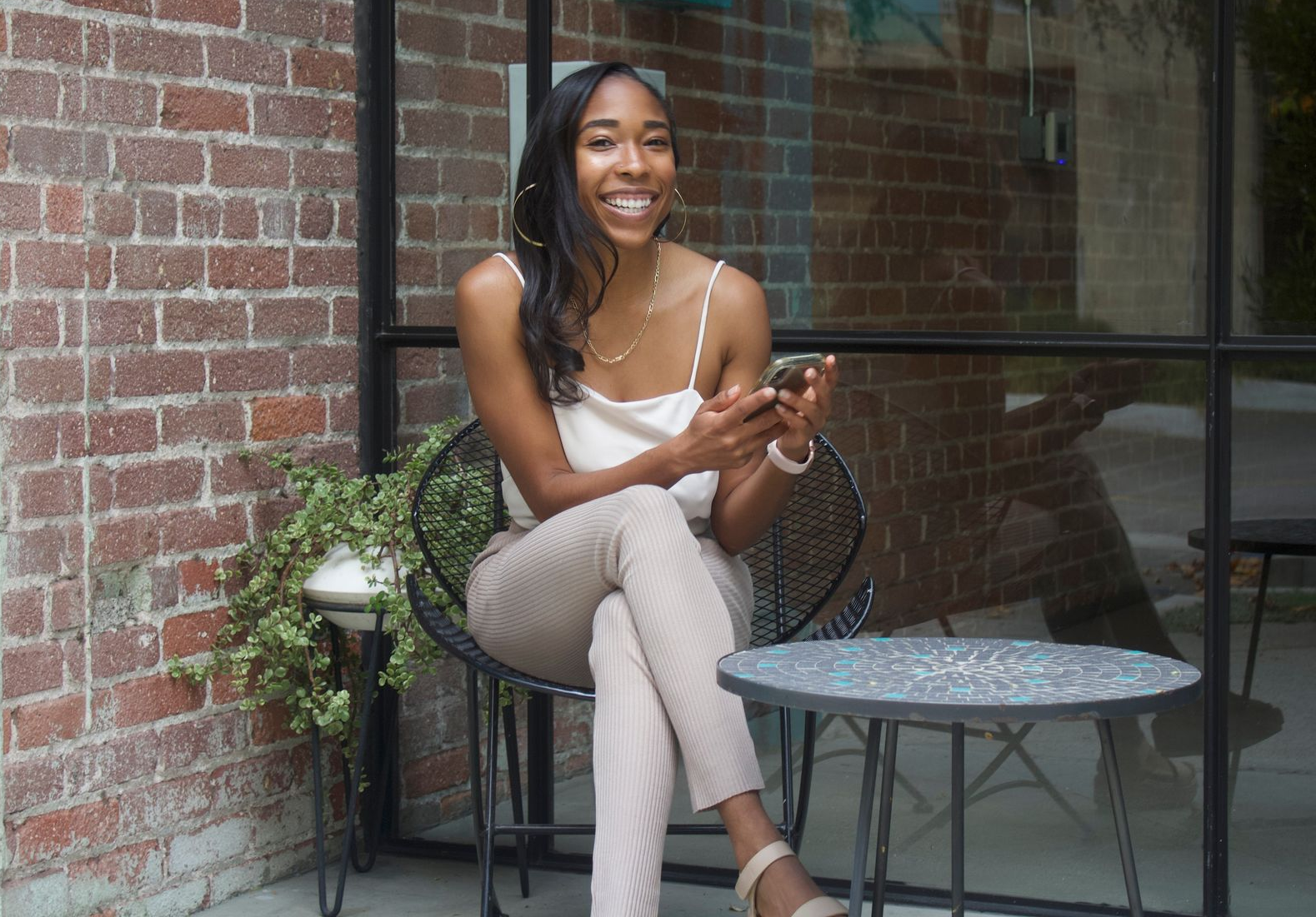 Woman sitting on a patio chair beside a small table, smiling in front of a brick wall.