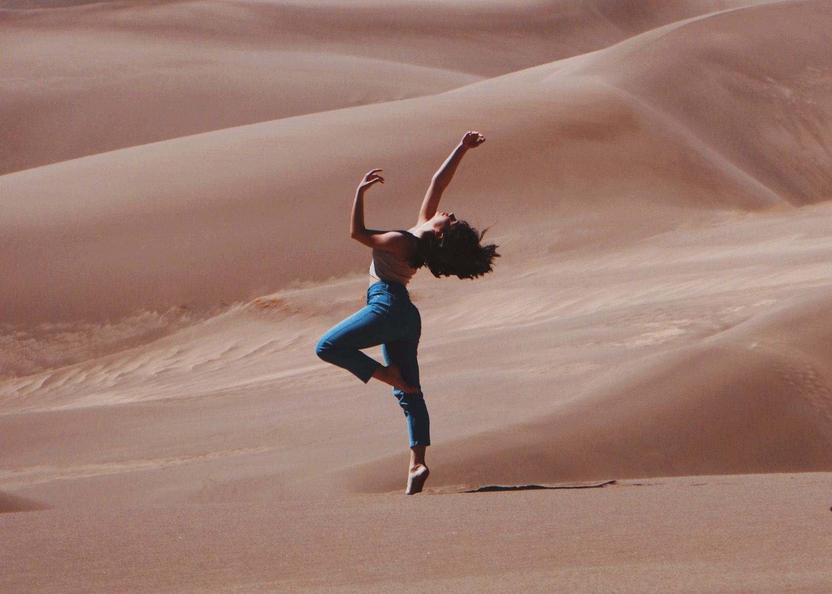 Person dancing on one foot in blue pants on sand dunes under a clear sky