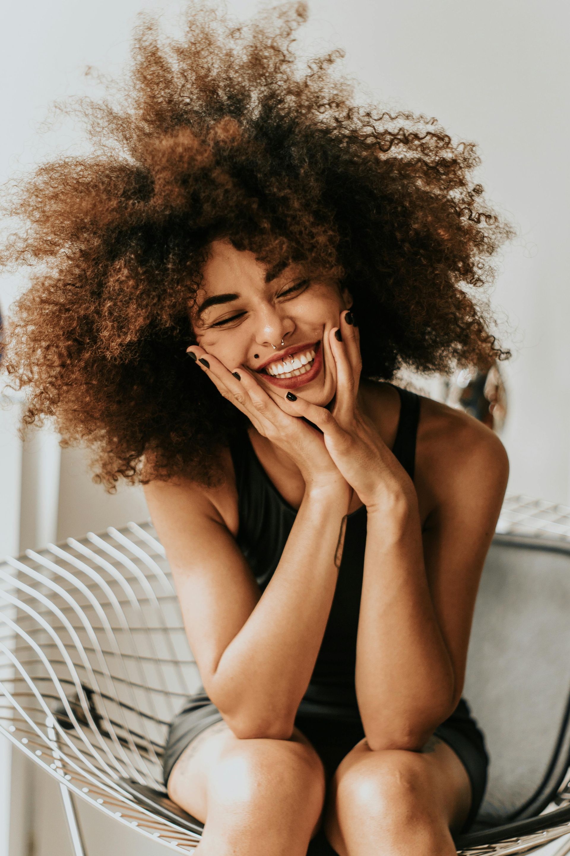 Smiling person with curly hair sitting on a white chair, resting their face in both hands.