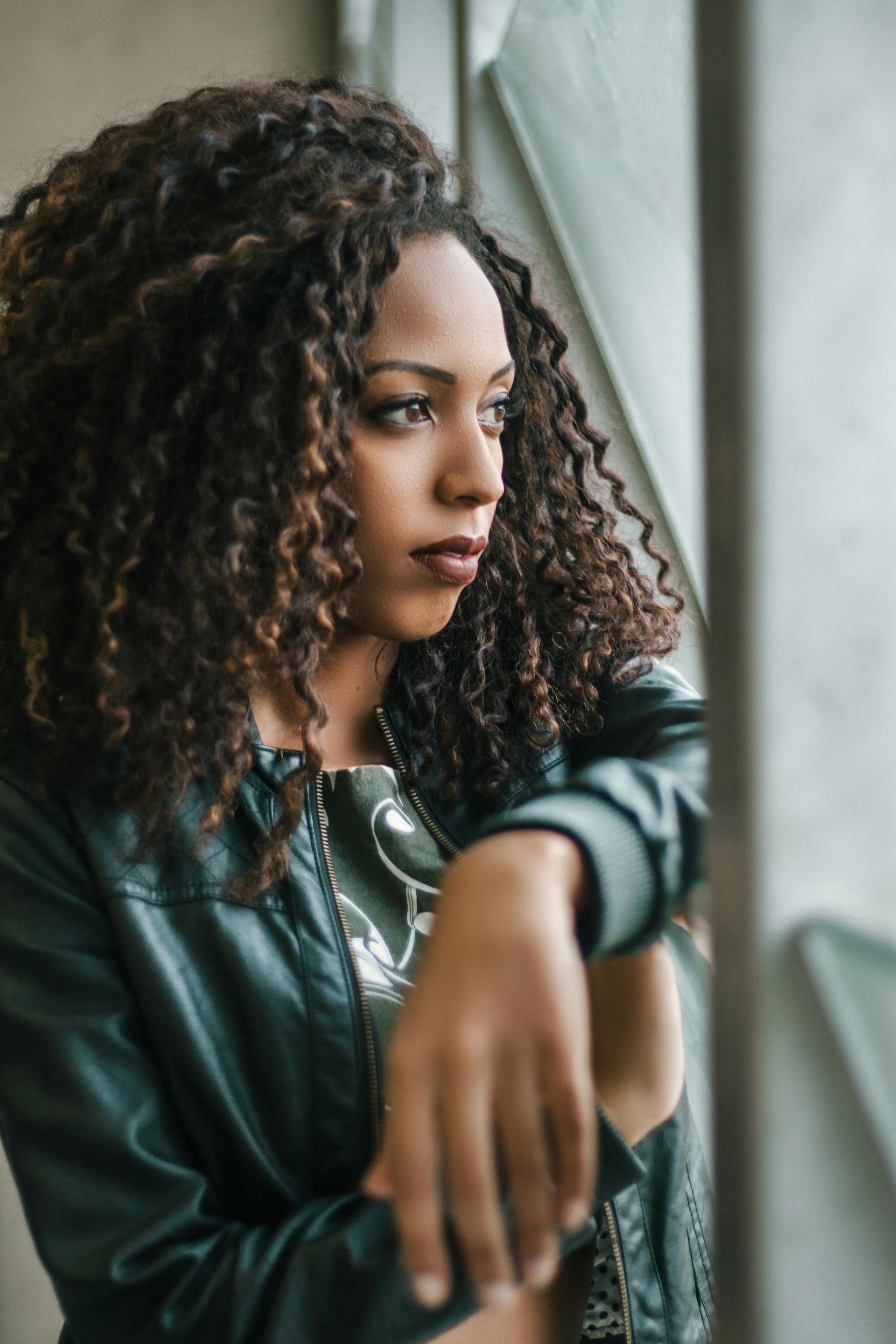 Person with curly hair leans by a window, gazing outside in a dark jacket.