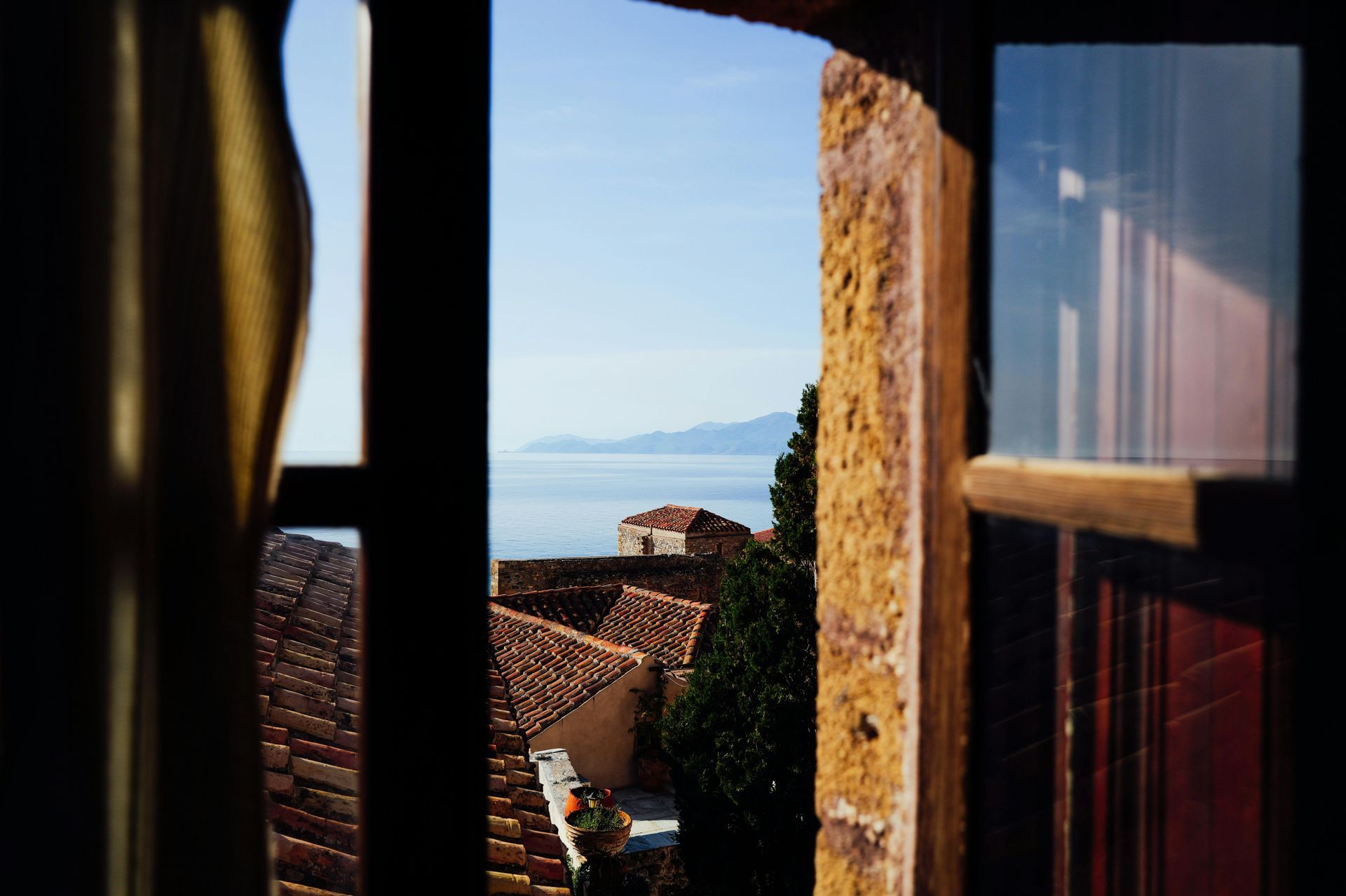 View through a window to red-roofed buildings, blue sea, and distant mountains in warm sunlight
