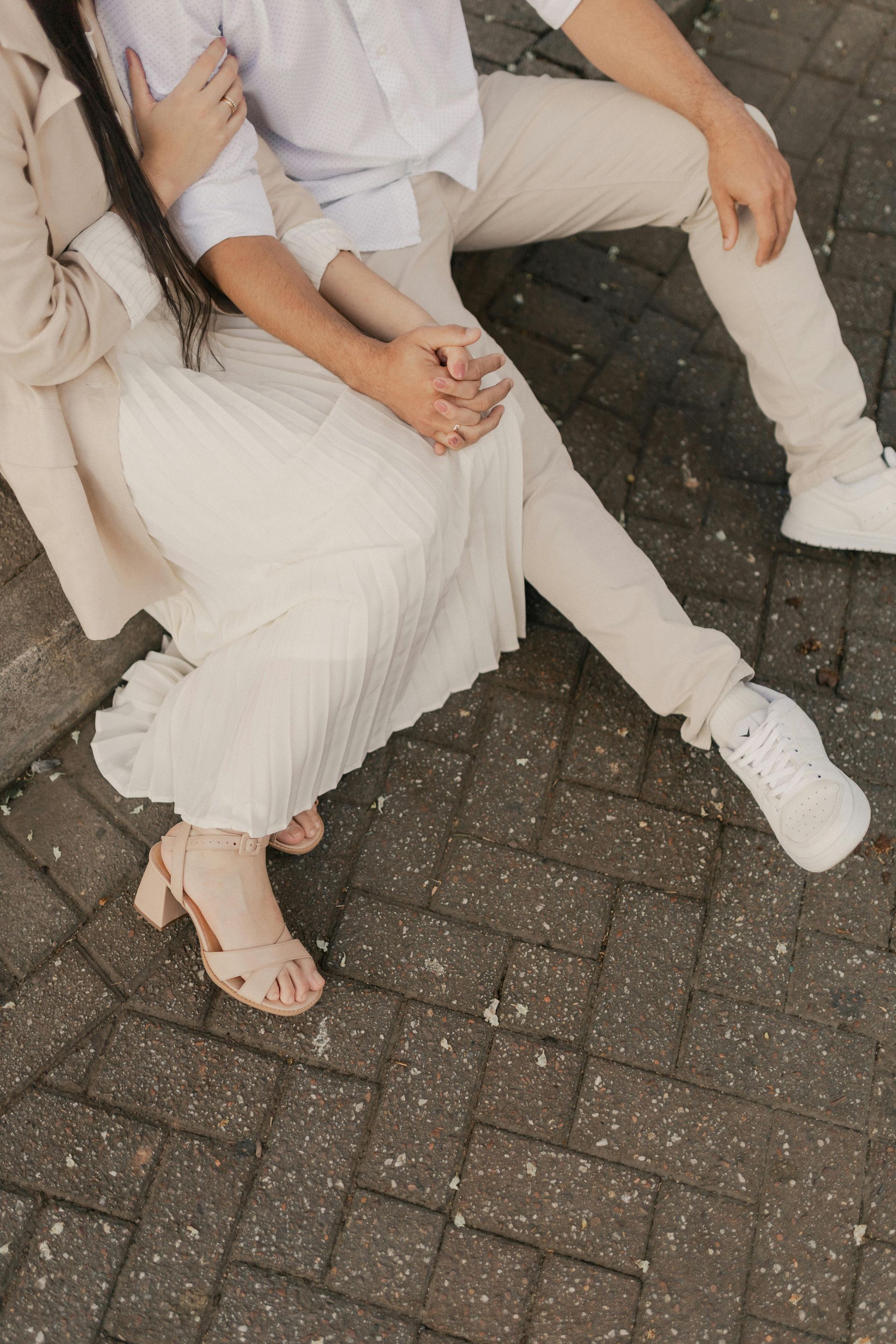 Two people sitting close together on a paved path, wearing light-colored clothes and holding hands.
