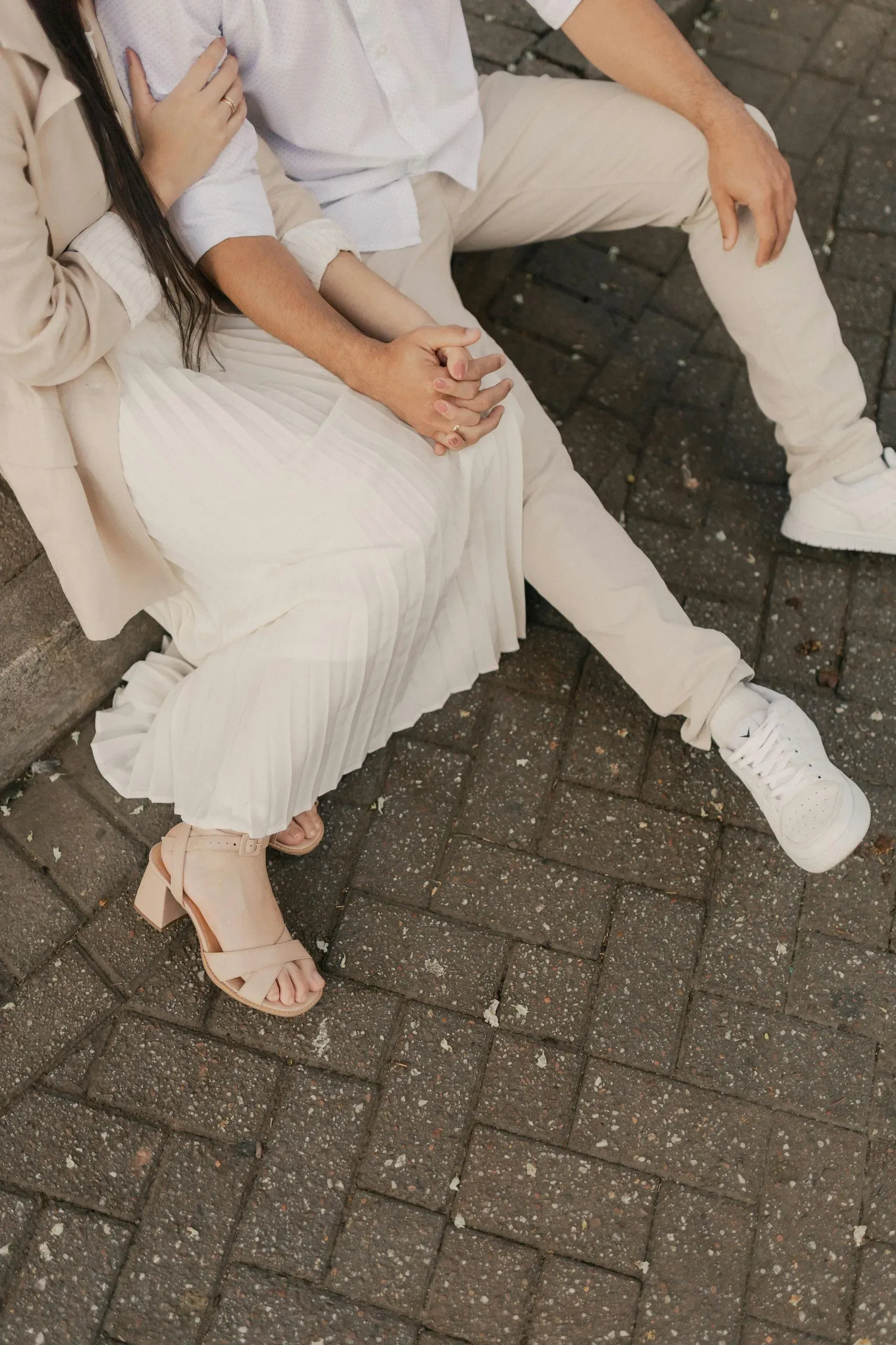 Two people sitting close together on a paved sidewalk, wearing light-colored clothes and white sneakers.