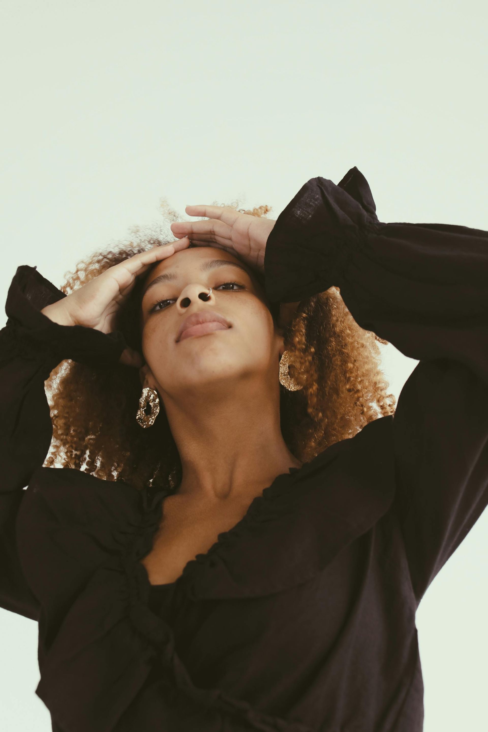 Person in a black top posing with hands in hair against a light background