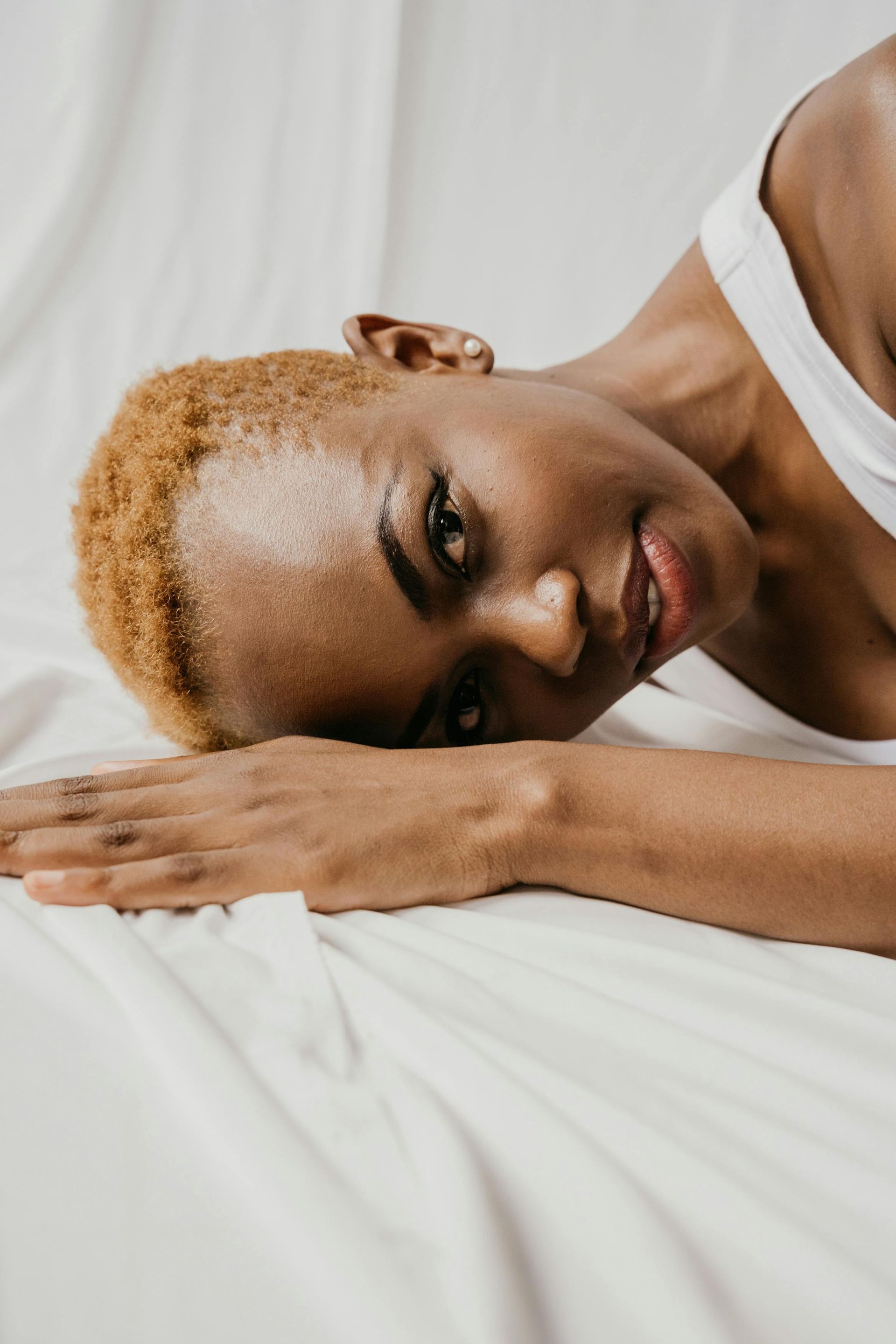 Person with short blond hair lying on a white pillow, resting their head on folded hands, looking at the camera.