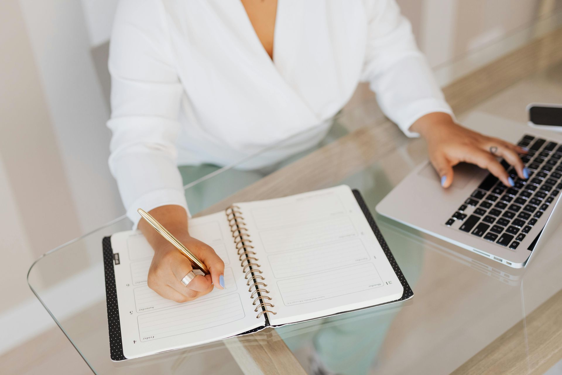 Person writing in a notebook beside a laptop on a glass desk