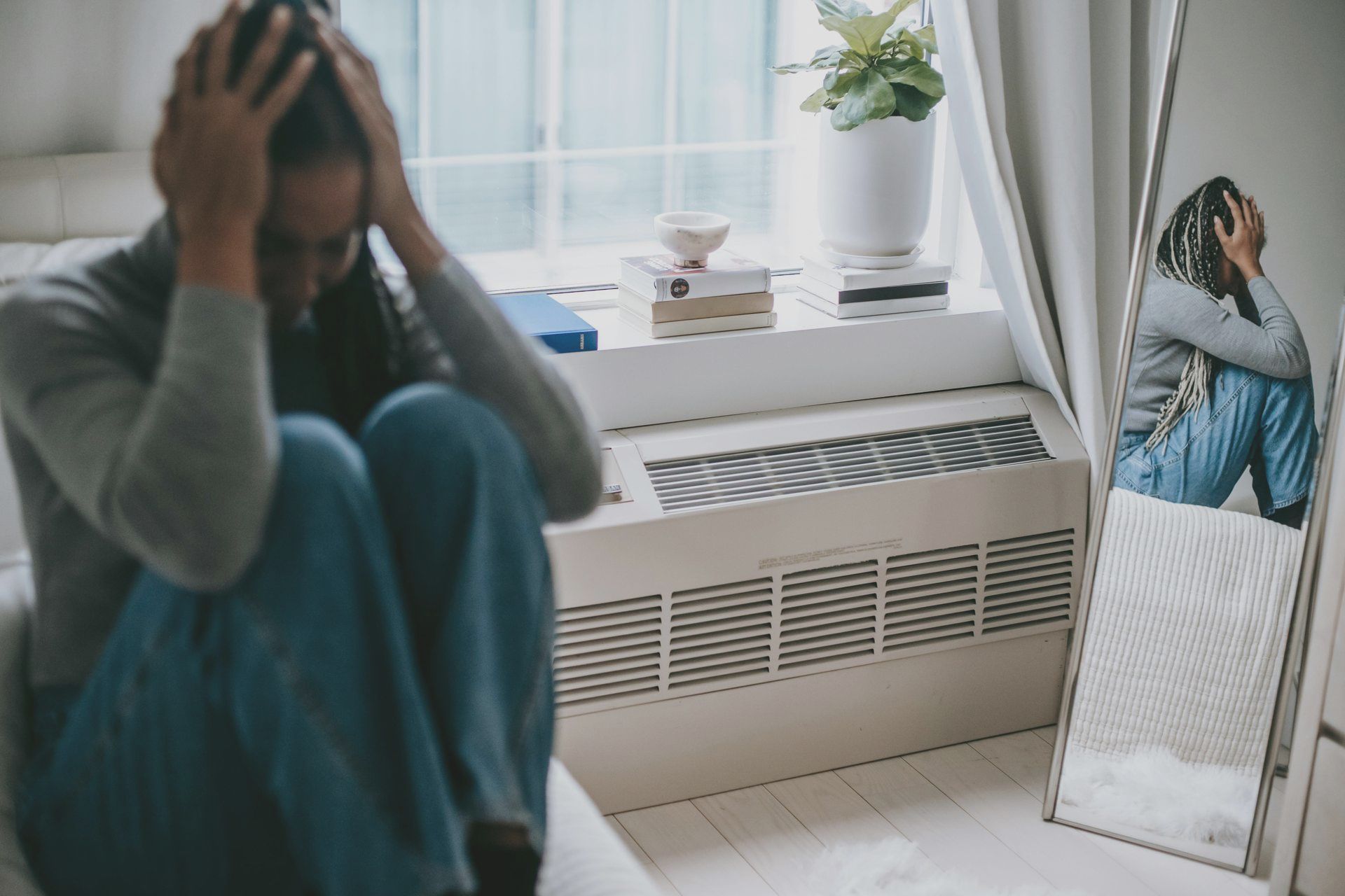 Person sitting on a bed with hands on head, reflected in a mirror beside a window and radiator.