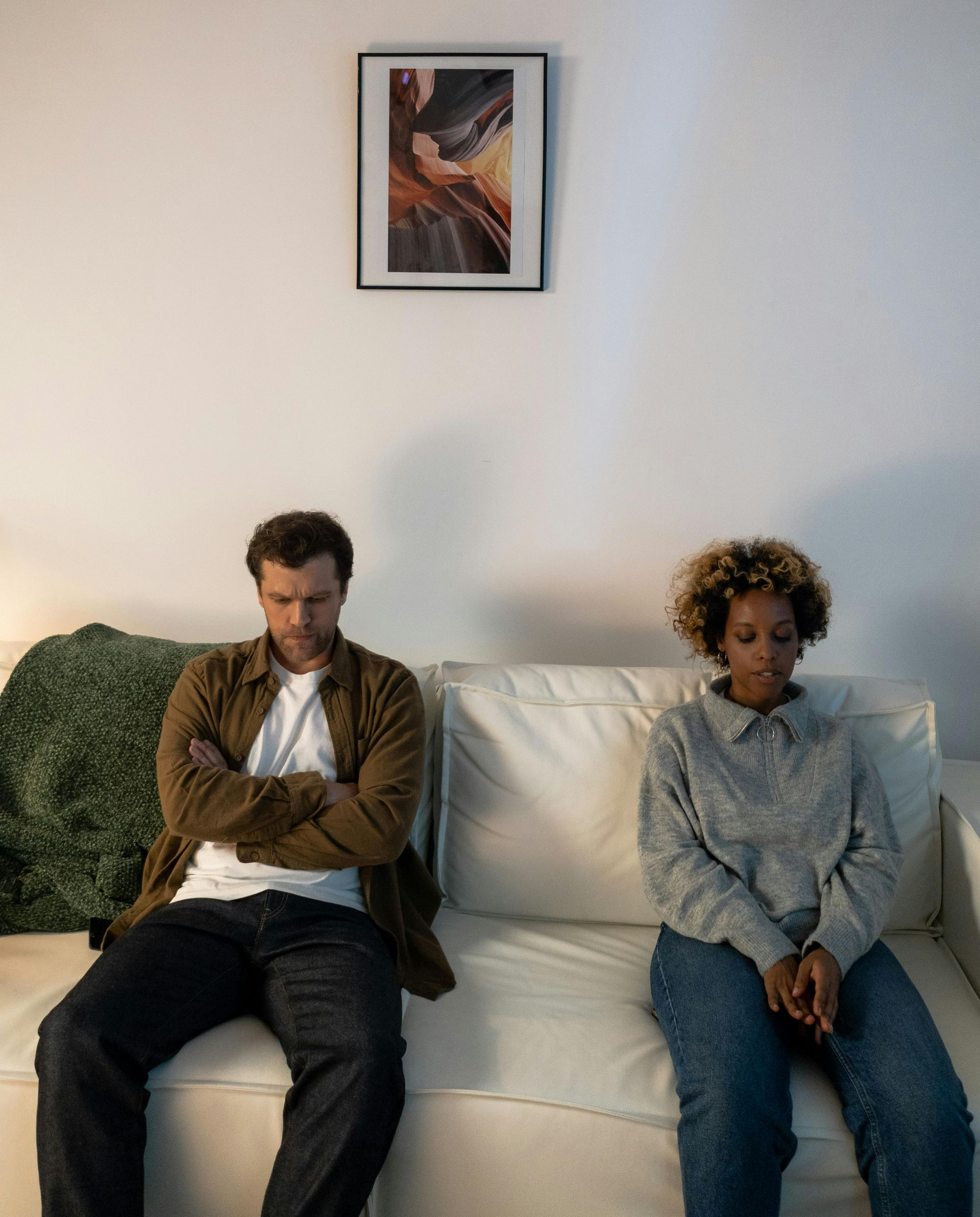 Two people sit apart on a white couch in a sparse living room, both looking down with serious expressions.
