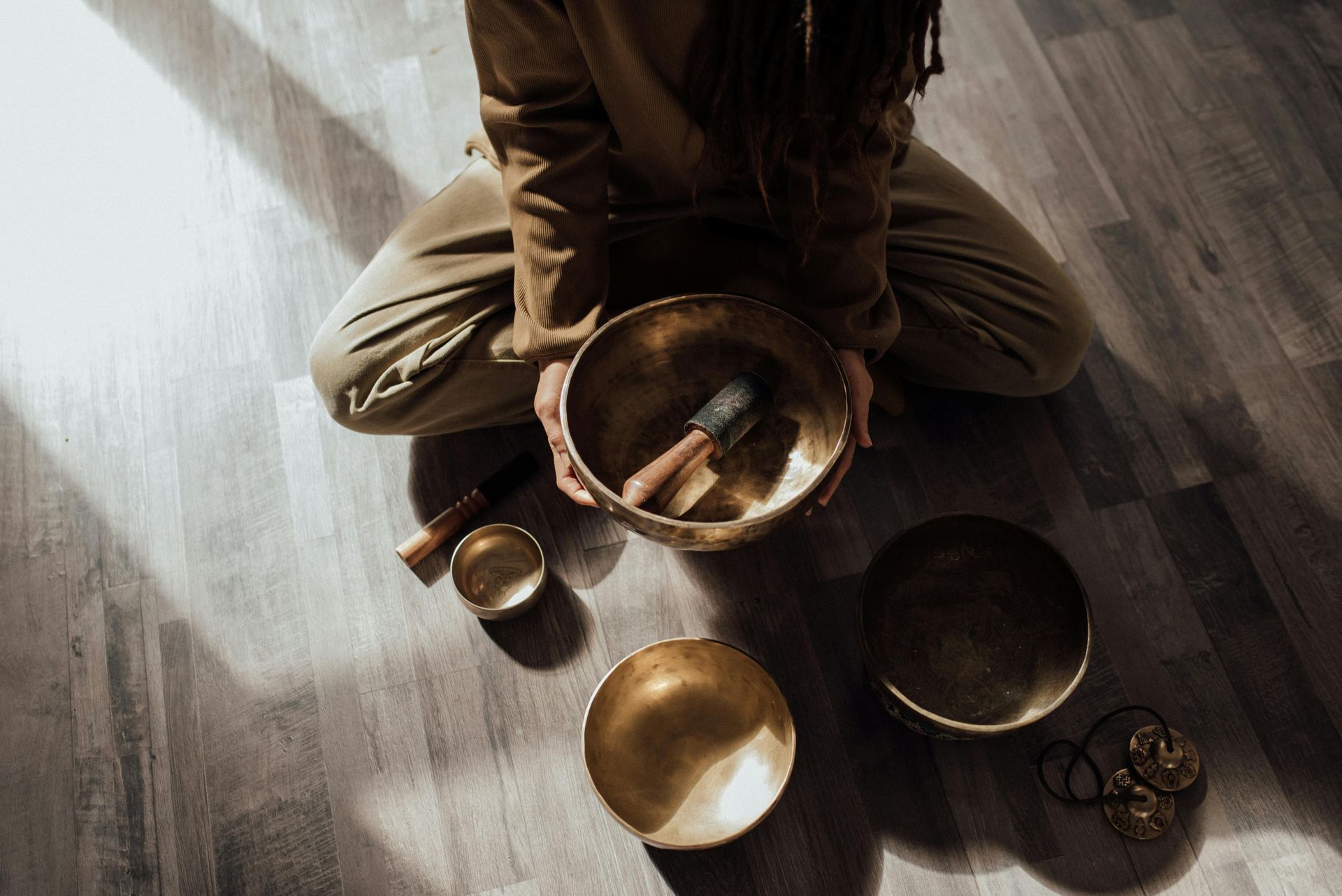 Person seated on a wooden floor playing a set of brass singing bowls in warm sunlight