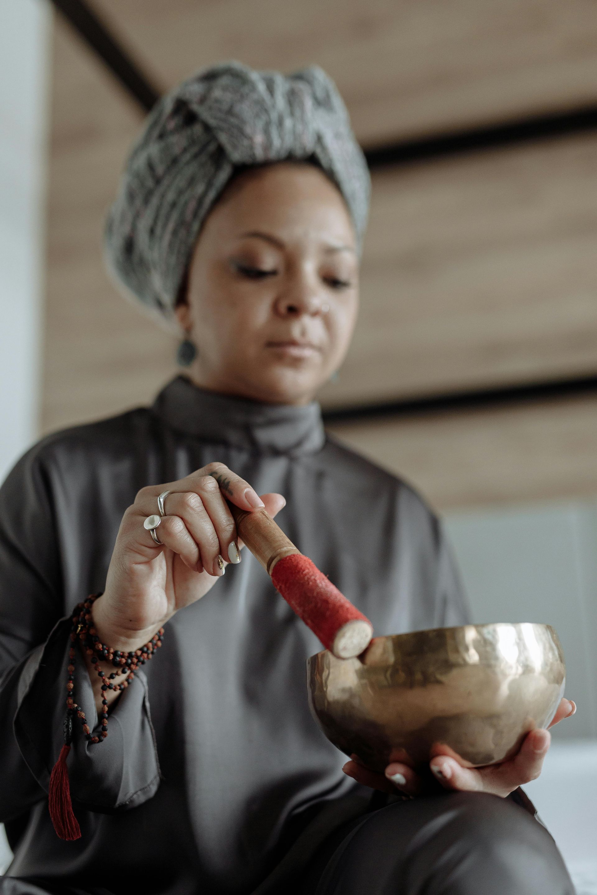Person holding a singing bowl and using a red mallet, wearing a gray robe and headwrap.