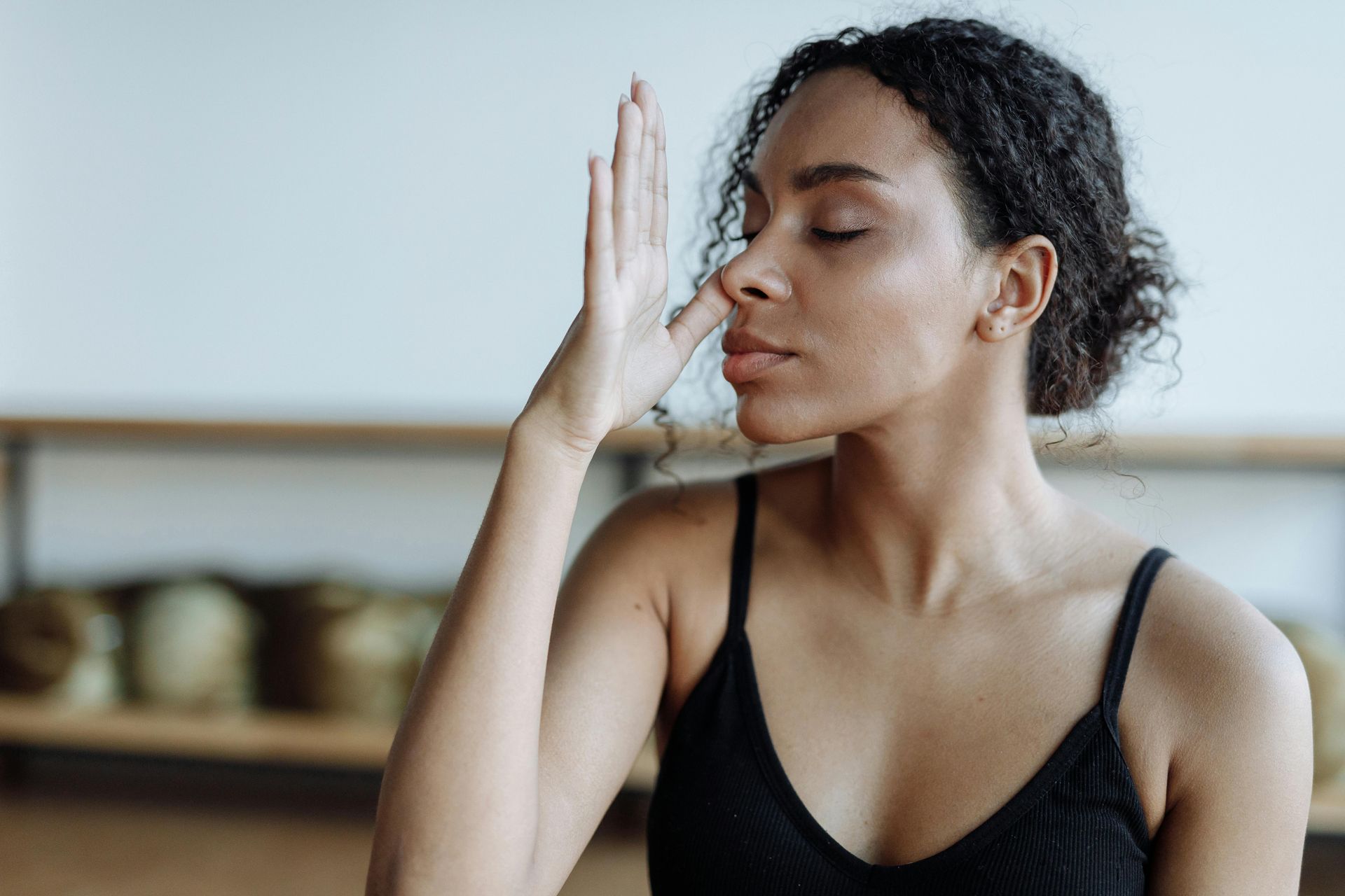 Person in a black tank top sits indoors with eyes closed, holding a hand to their face in a meditative pose.