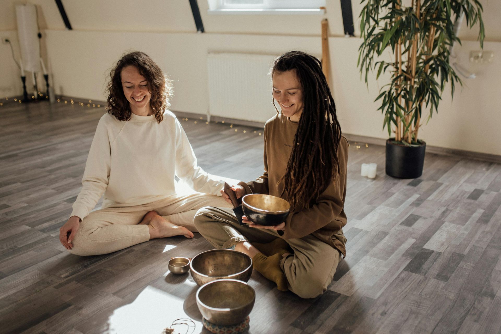 Two people seated on a wooden floor with singing bowls, meditating in a bright room
