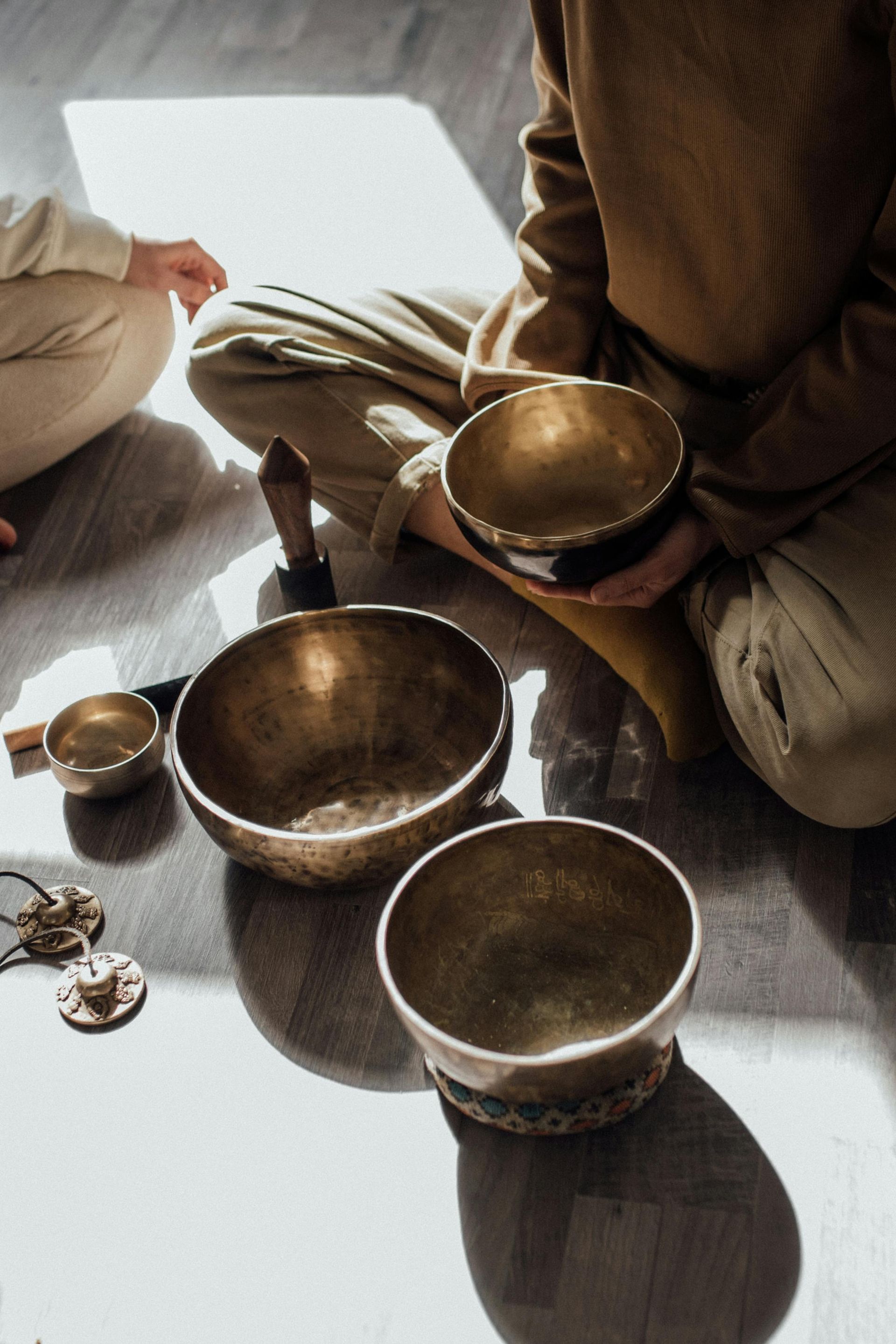Two people sit on the floor with singing bowls in soft sunlight, one holding a bowl and mallet.