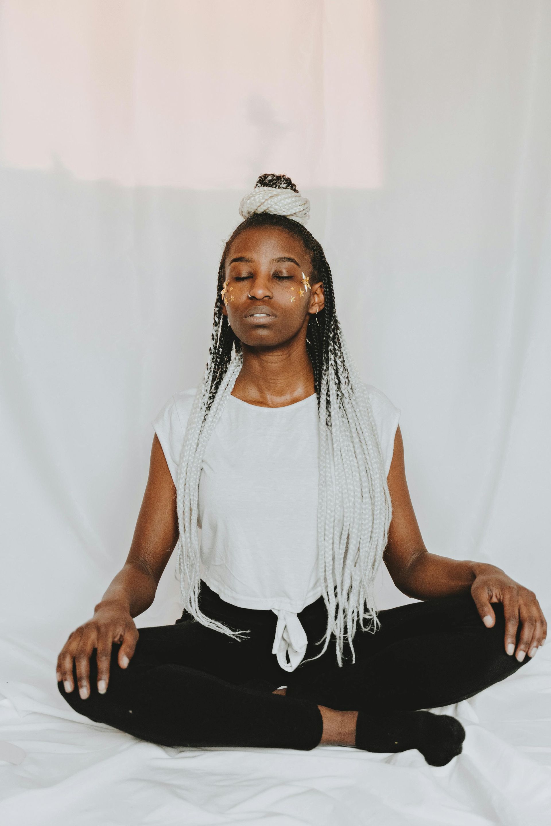 Person sitting cross-legged on a white sheet, with long white braided hair, eyes closed, in a calm pose.