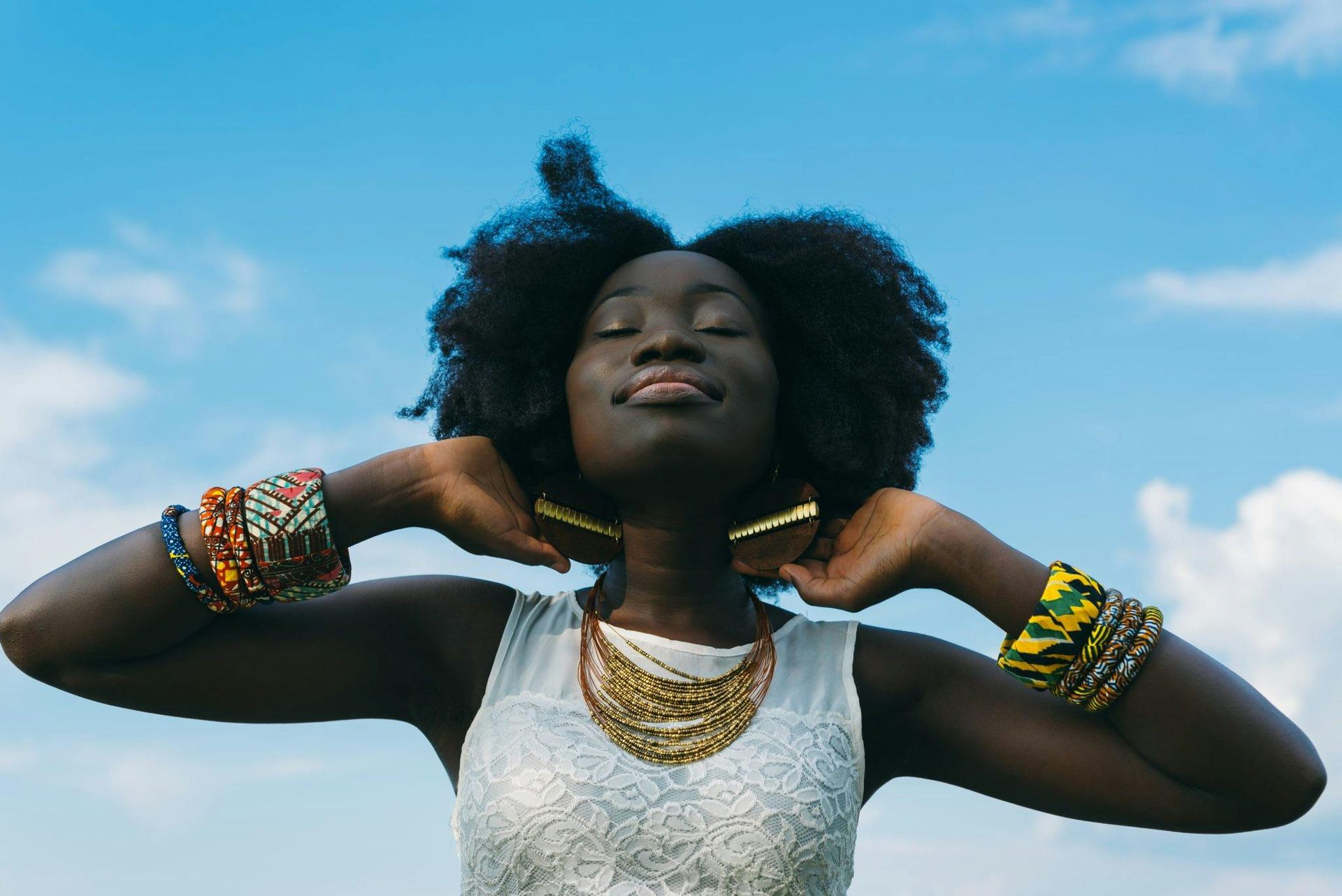 Woman in white top with gold jewelry, adjusting her necklace against a blue sky.