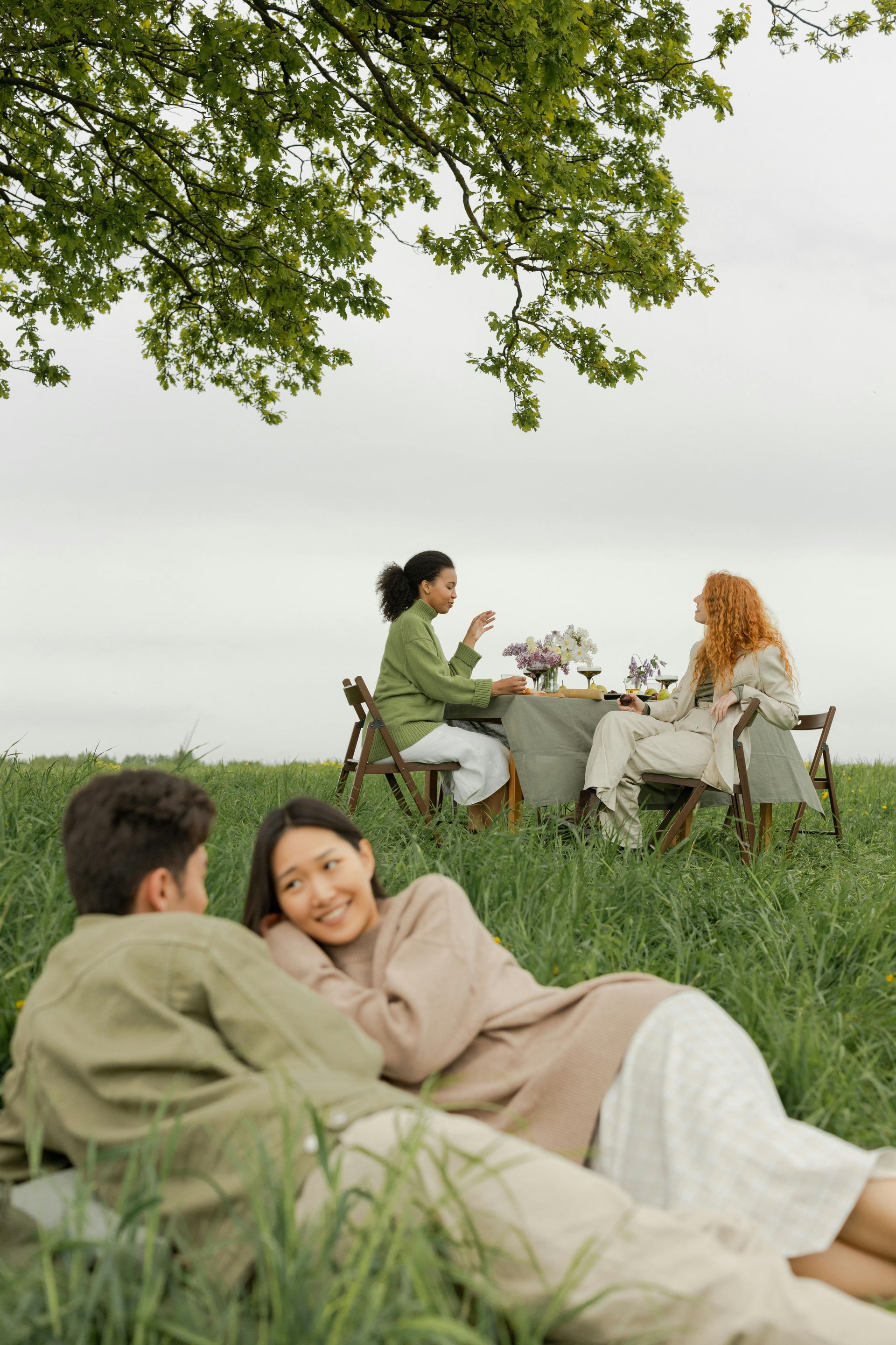 Two people relaxing on grass near a table, with two more seated in a grassy field under a tree.