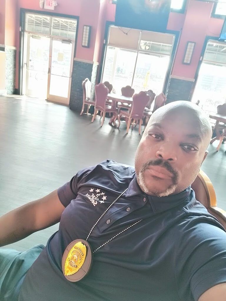 A Black man in a blue shirt with a badge, seated in a restaurant.
