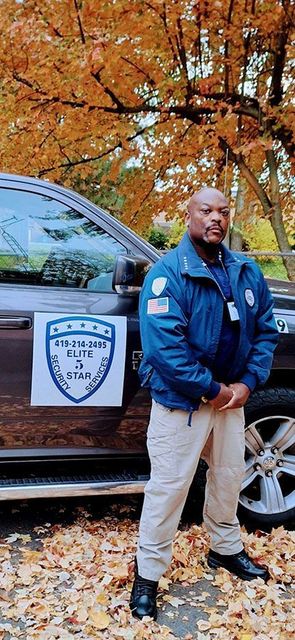 A man in blue jacket and tan pants stands by a car with fall foliage in the background.