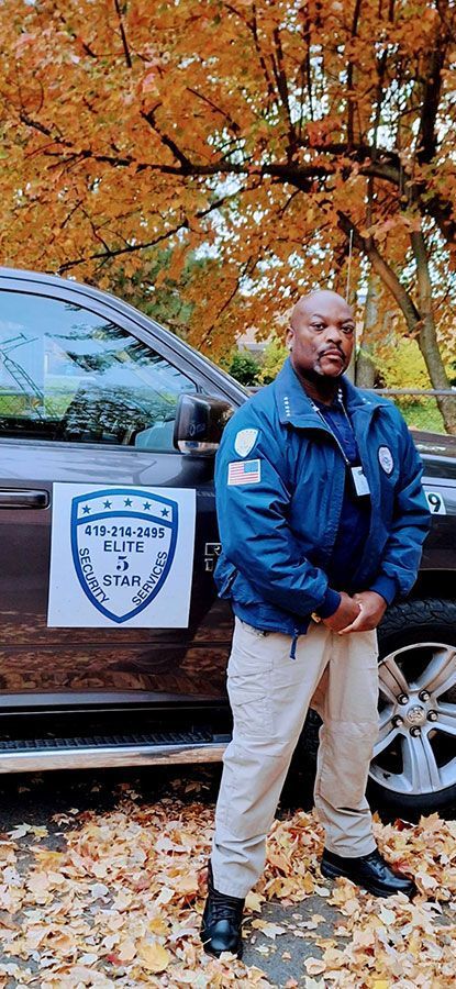 A Black man in uniform stands next to a vehicle with fall foliage in the background.