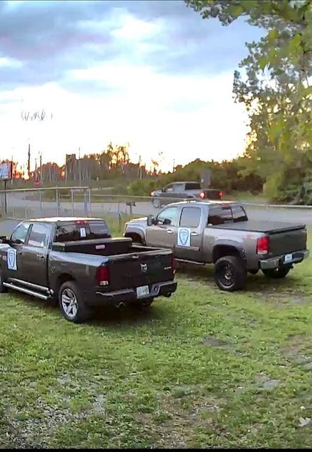 Three pickup trucks parked on grass. Gray and black trucks in a lot, with a fourth truck in the background.