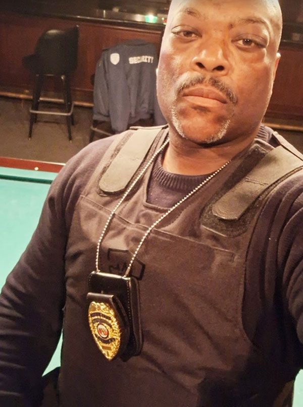 Black man in a security vest, badge visible.  He looks directly at the camera, indoors.