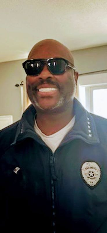 Man wearing sunglasses, smiling, in a dark jacket with a badge.