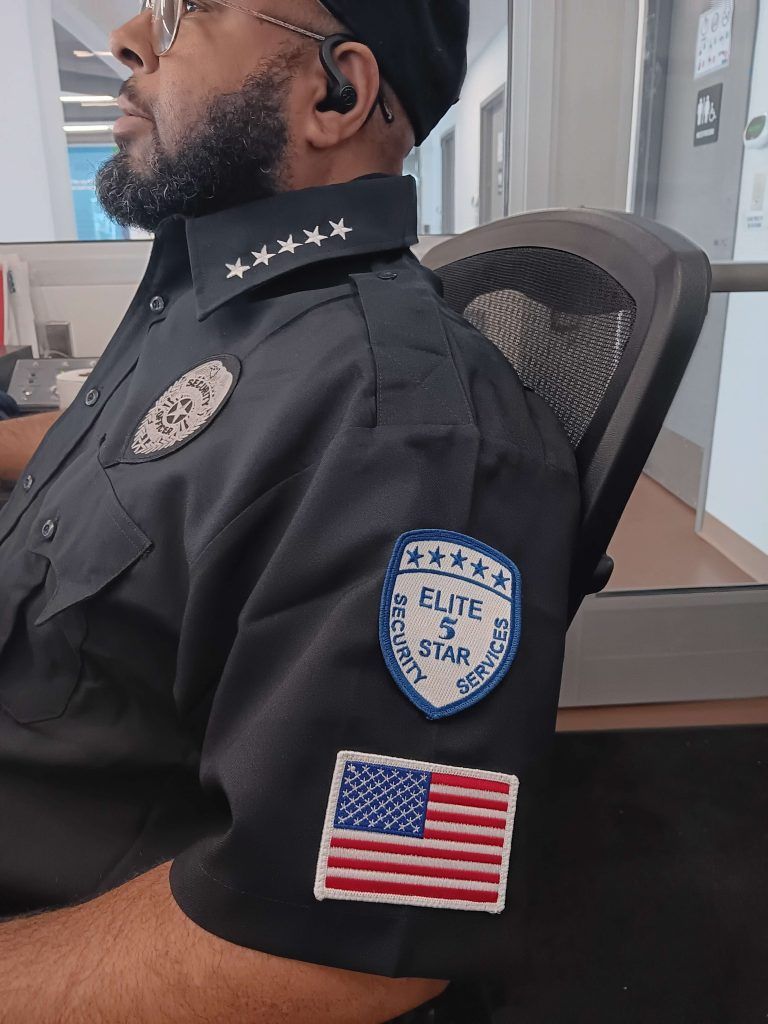 Man in security uniform with a badge and American flag patch, sitting indoors.