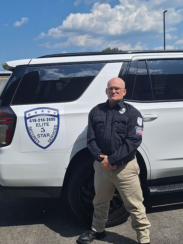 Man in uniform stands next to a white SUV with logo. He wears a dark blue shirt, khaki pants, and has short hair.