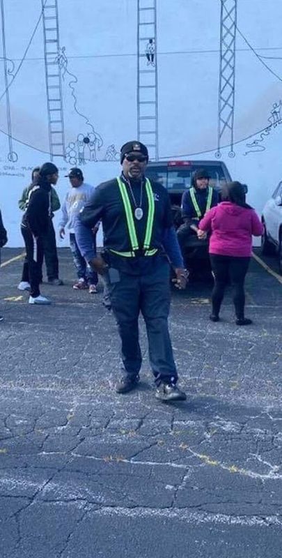 A man in uniform and sunglasses walks towards the camera in a parking lot with other people.