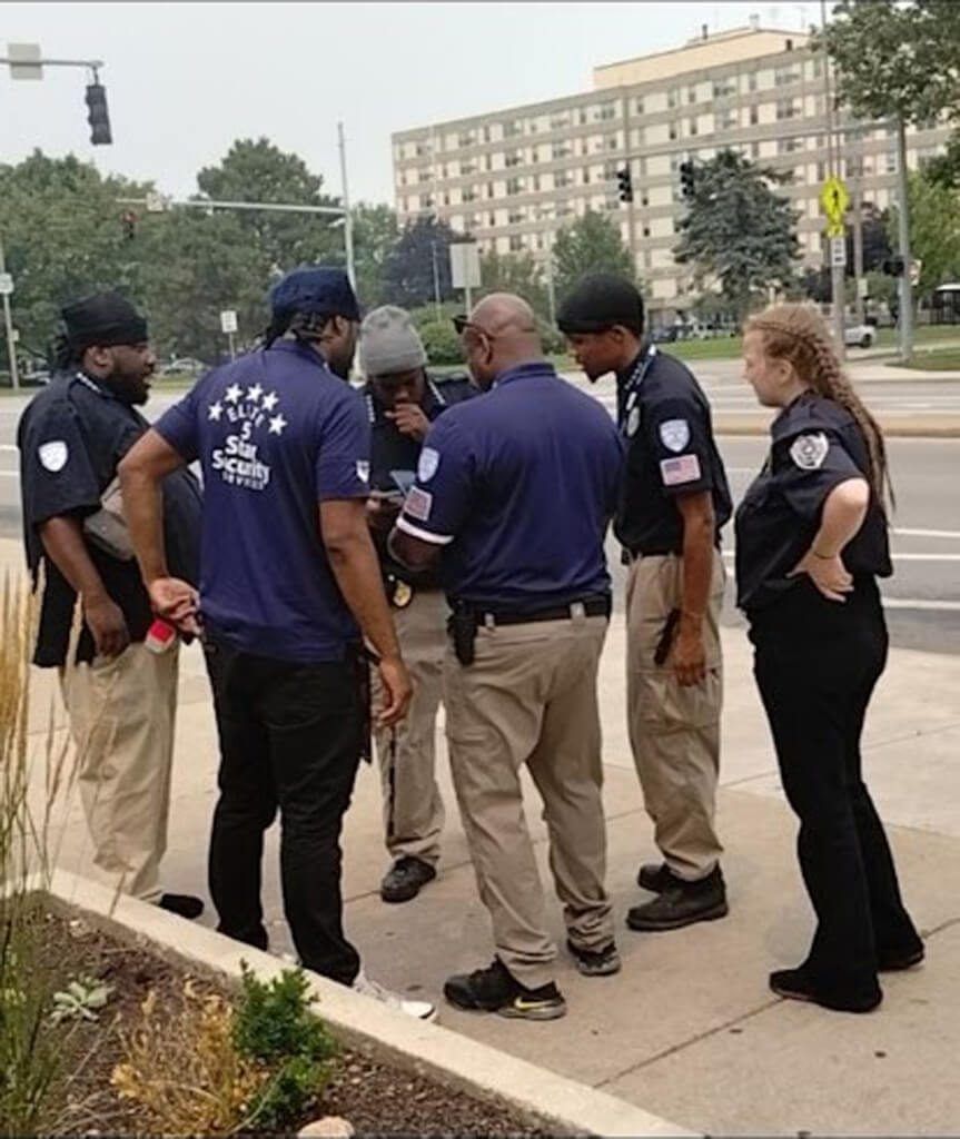 A group of security personnel stand on a sidewalk, looking at a phone. They are in front of a building.