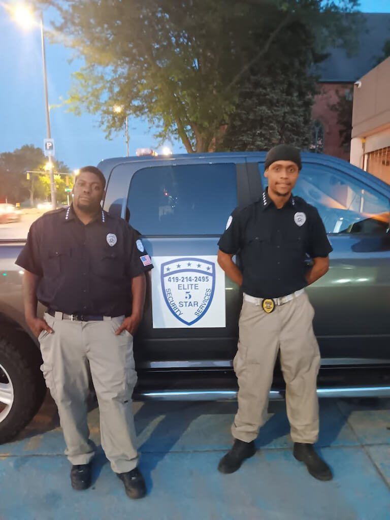 Two security guards pose next to a truck with a company logo. They wear dark shirts and tan pants outdoors.